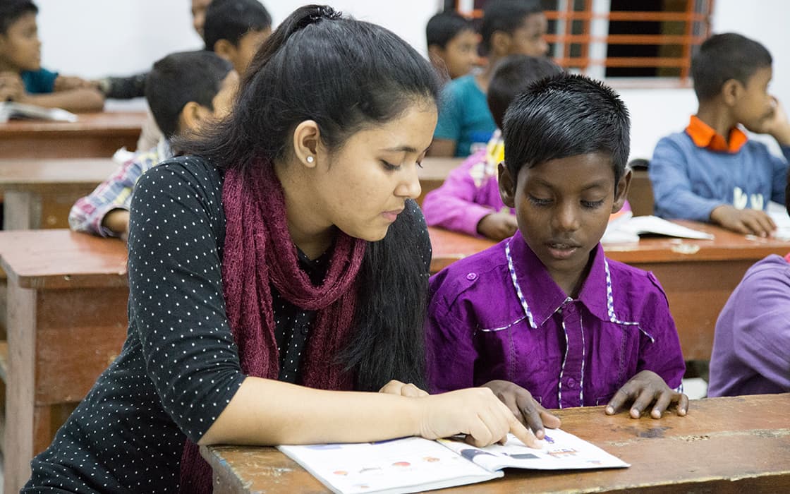 Ein Lehrer hilft einem Jungen beim Lesen eines Buches in einem Klassenzimmer, in dem andere Schüler an Holzpulten sitzen.