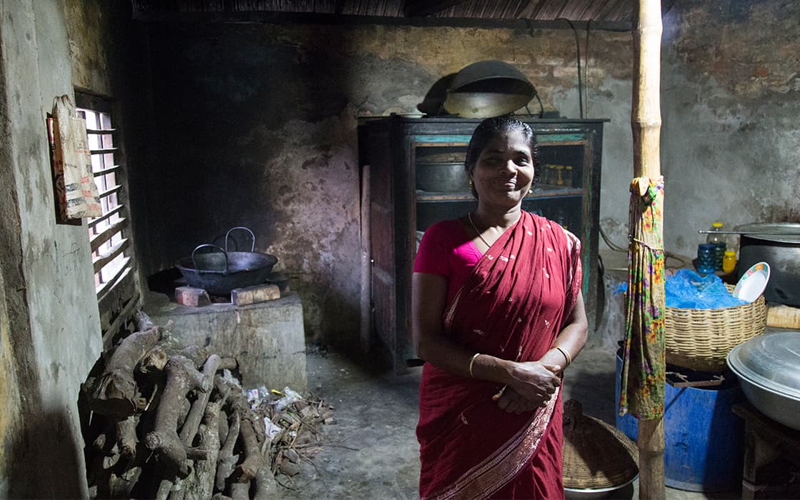 Eine Frau in einem roten Sari steht lächelnd in einer rustikalen Küche mit gestapeltem Feuerholz und Kochutensilien um sie herum.