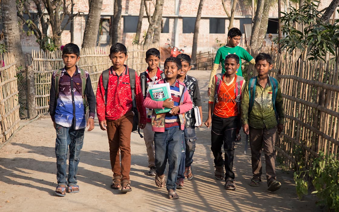 Eine Gruppe von Jungen mit Rucksäcken und Büchern geht gemeinsam auf einem sonnigen Weg, der von Bambuszäunen und Bäumen gesäumt ist.