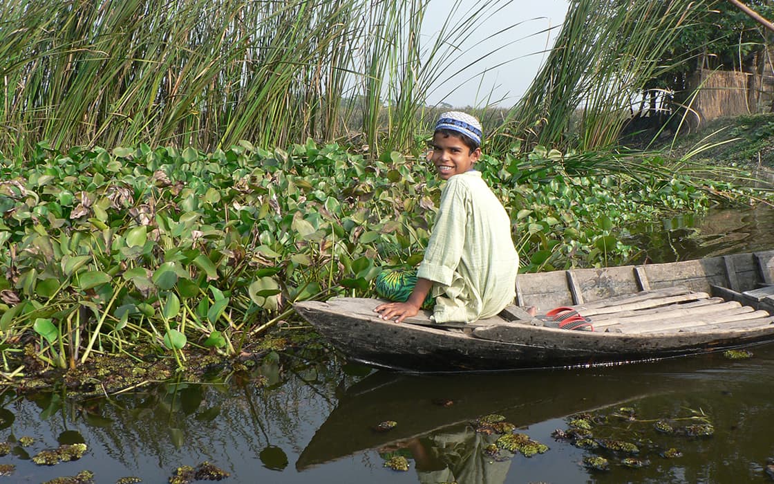 Ein Junge in einem Boot lächelt, während er in der Nähe von hohem Schilf und Wasserpflanzen auf einem ruhigen Fluss sitzt.