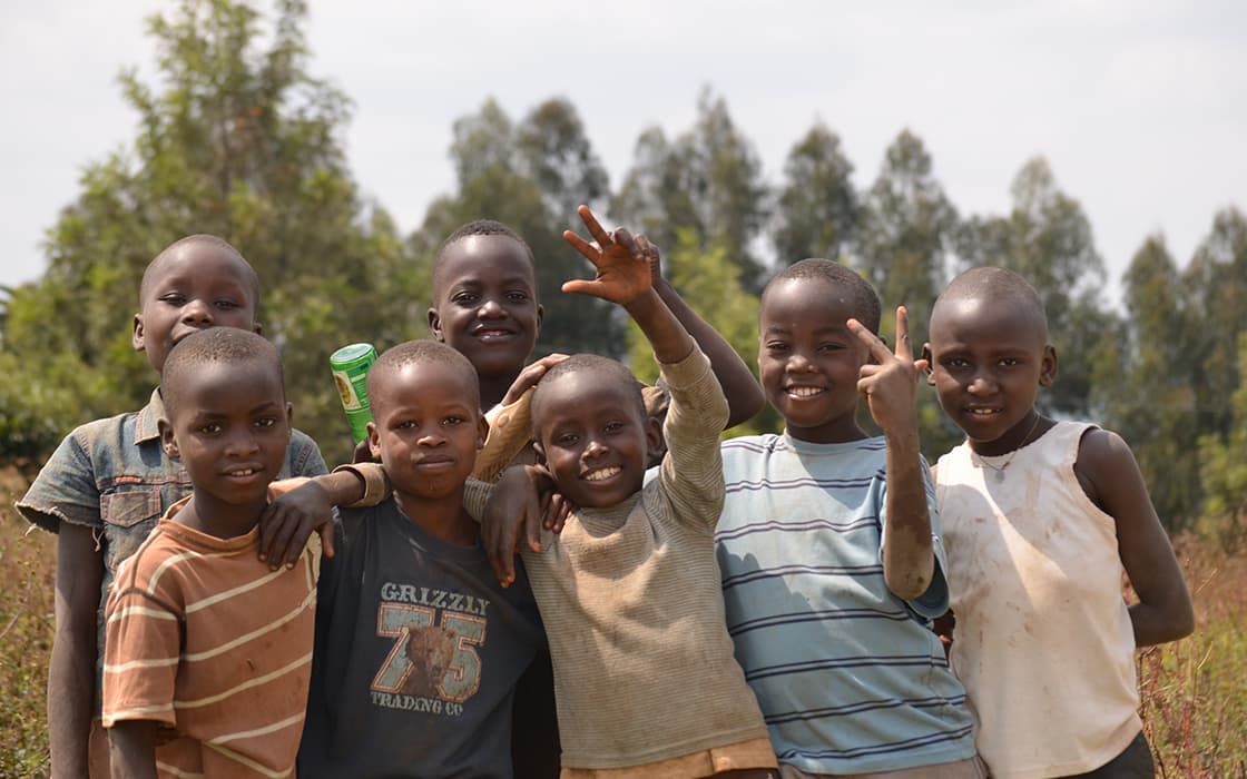 Sieben lächelnde Kinder stehen im Freien vor Bäumen, einige machen Friedenszeichen, andere legen die Arme umeinander.