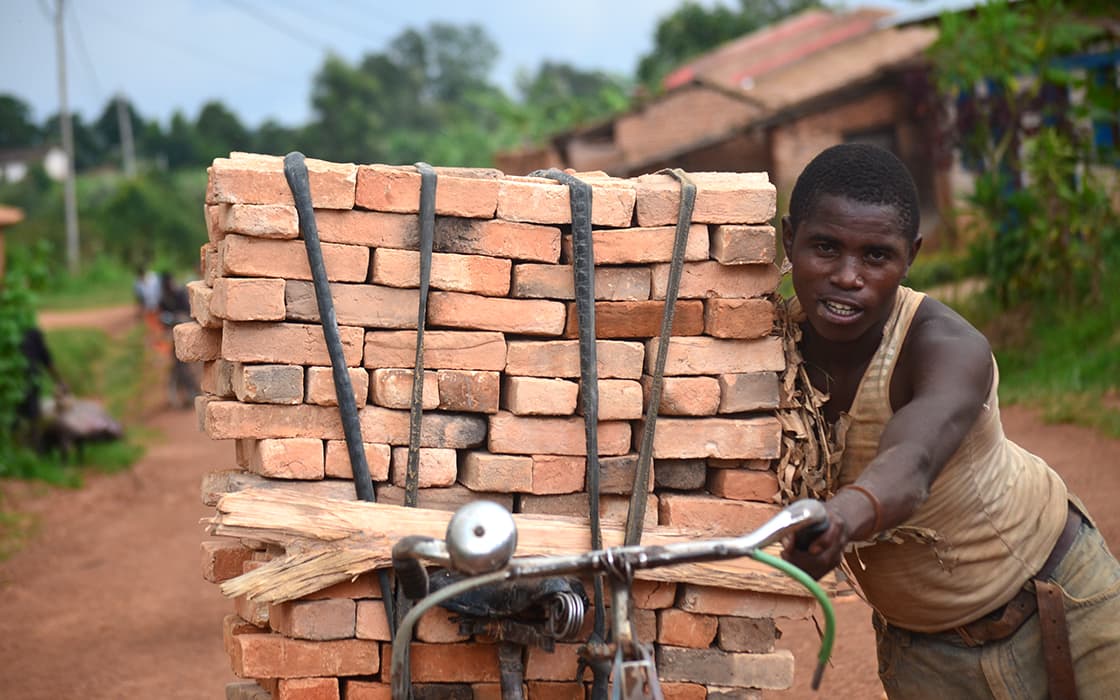 Eine Person schiebt ein mit einem großen Stapel Ziegelsteine beladenes Fahrrad über einen Feldweg in einer ländlichen Gegend.