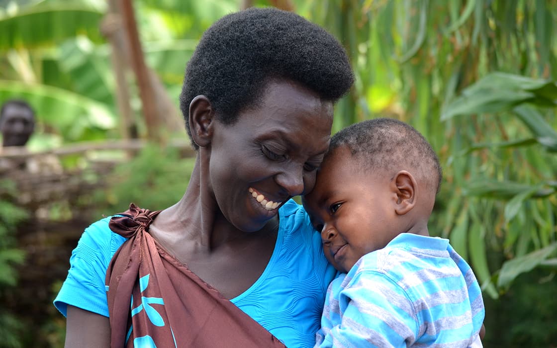 Eine lächelnde Frau umarmt ein kleines Kind im Freien, umgeben von grünen Pflanzen.