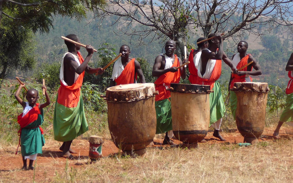 Eine Gruppe von bunt gekleideten Menschen spielt im Freien auf großen Trommeln, während ein Kind daneben tanzt.