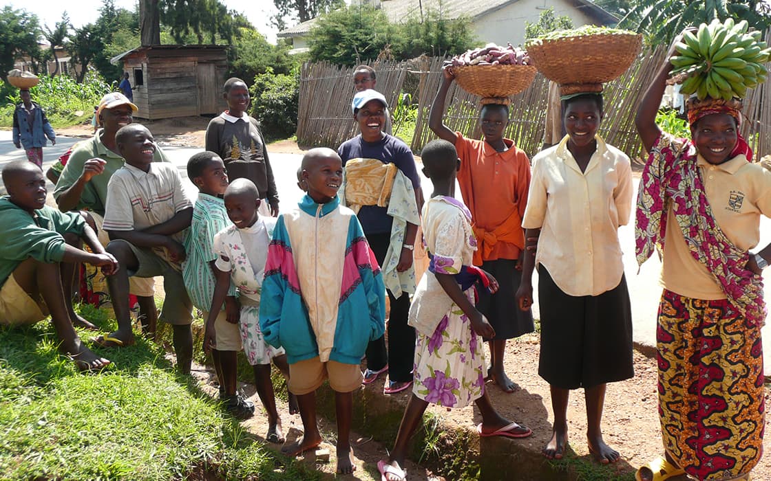 Eine Gruppe lächelnder Menschen, darunter Kinder und Frauen mit Körben auf dem Kopf, steht im Freien in einer Dorfumgebung.