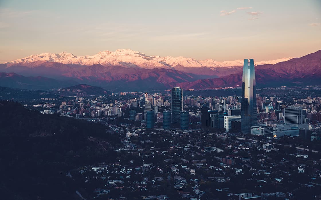 Stadtlandschaft mit hohen modernen Gebäuden, schneebedeckten Bergen im Hintergrund und einem rosafarbenen Himmel bei Sonnenuntergang.