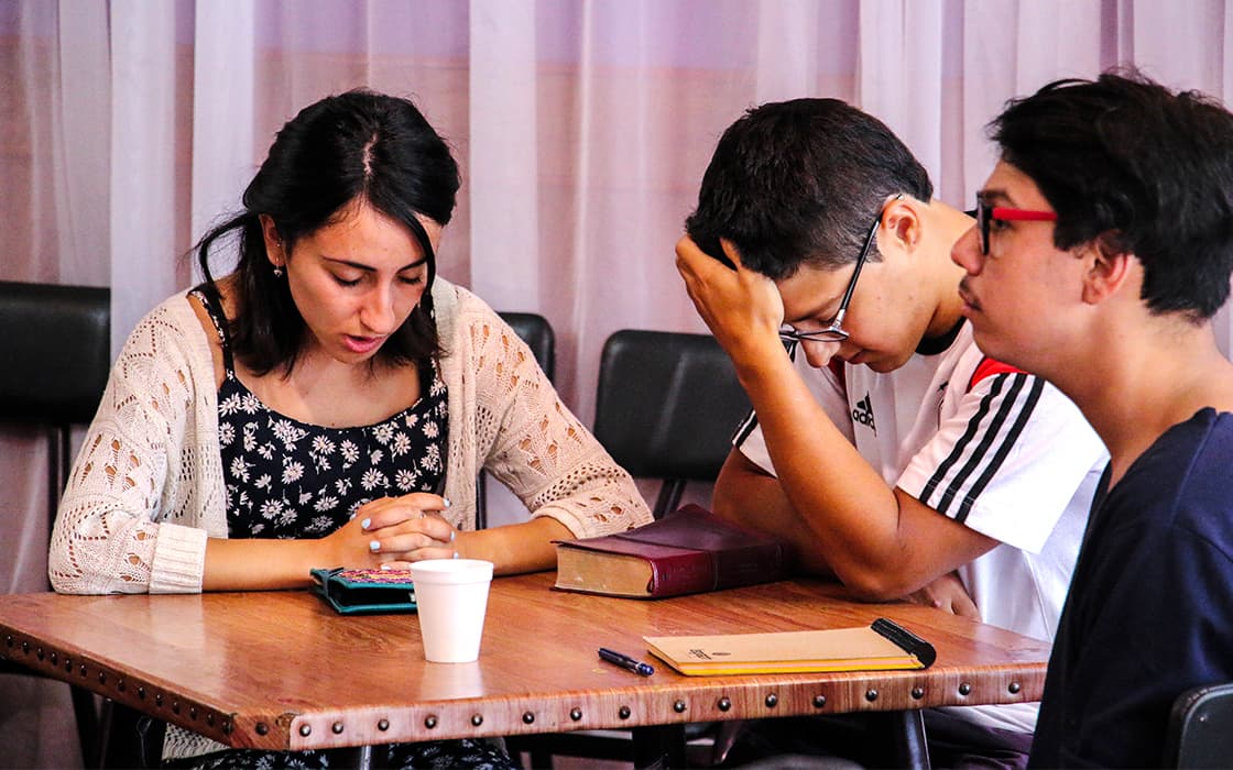 Drei junge Leute sitzen an einem Tisch, die Köpfe zum Gebet gesenkt, mit einem Buch, einem Notizbuch und einer Tasse vor sich.