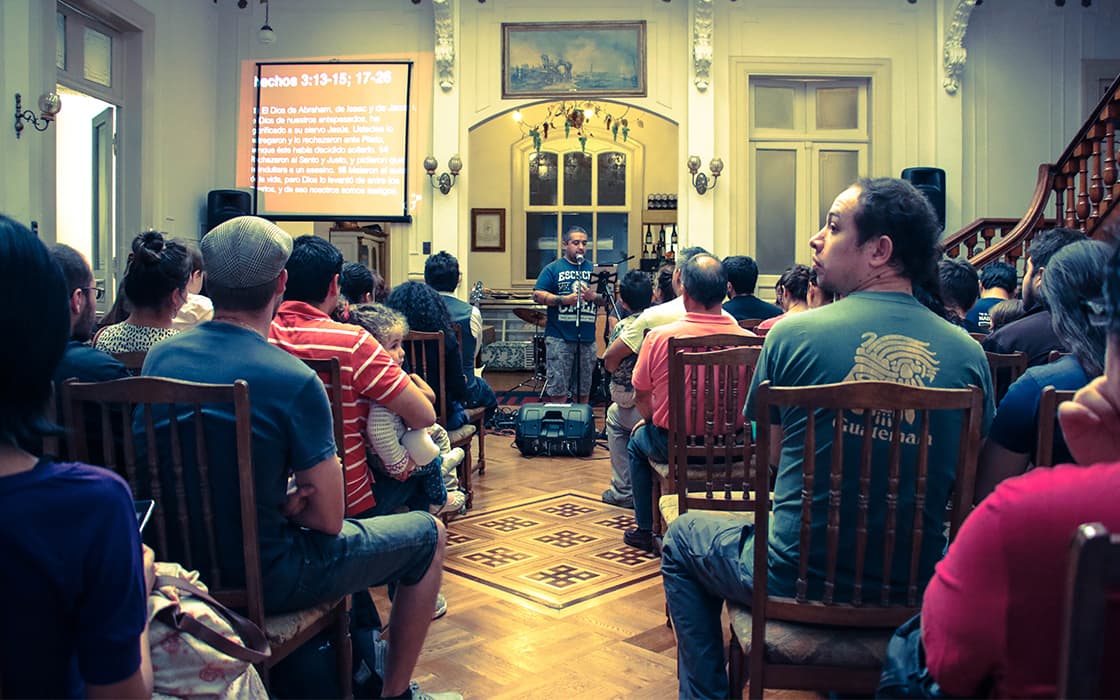 Ein Mann spricht zu einem Publikum in einem Raum mit Holzstühlen; hinter ihm wird ein Bibelvers auf eine Leinwand projiziert.