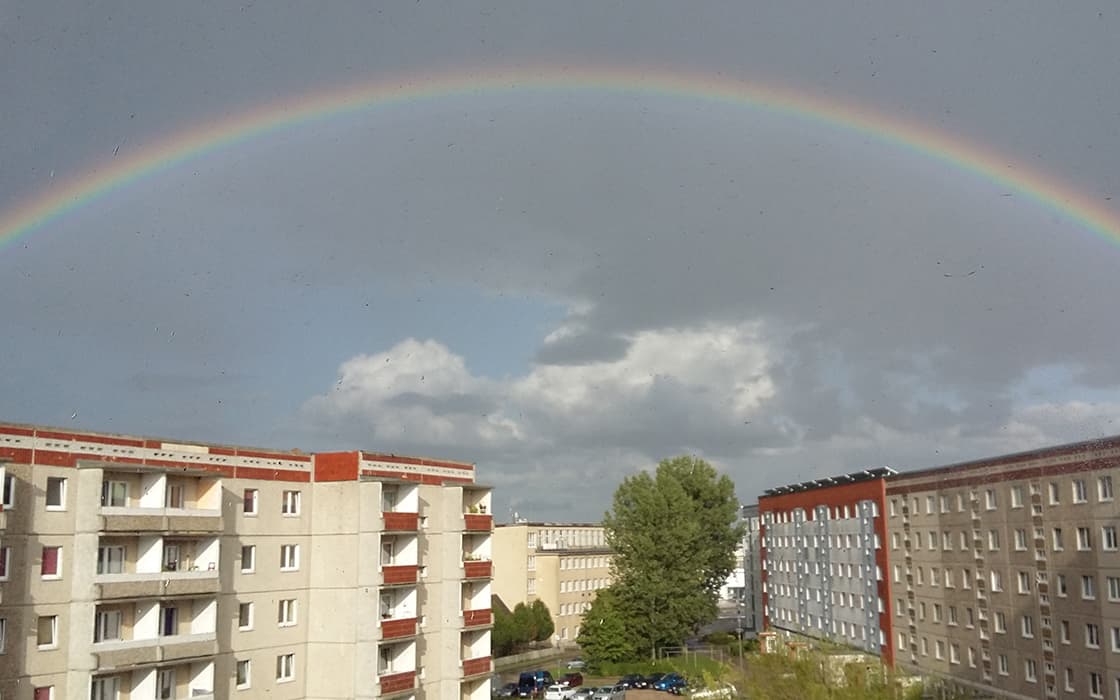 Ein Regenbogen spannt sich unter einem wolkenverhangenen Himmel über Wohnhäuser, unter denen Bäume und geparkte Autos stehen.