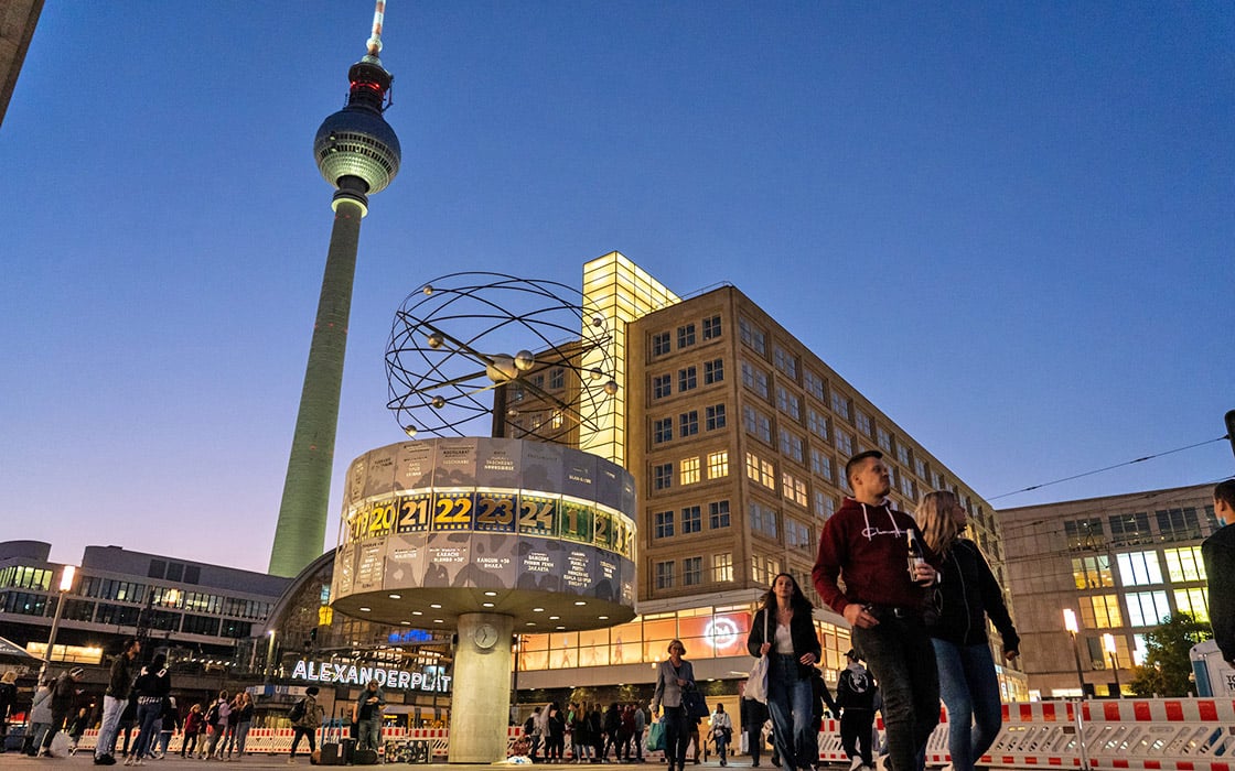 Spaziergänger auf dem Alexanderplatz in Berlin, mit Blick auf die Weltzeituhr und den Fernsehturm in der Abenddämmerung.