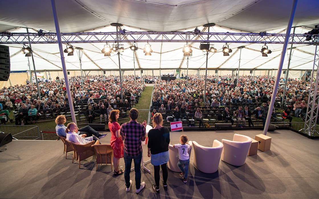 Blick von einer Bühne, auf der die Redner vor einem großen, sitzenden Publikum in einem hellen, überdachten Veranstaltungsraum stehen.