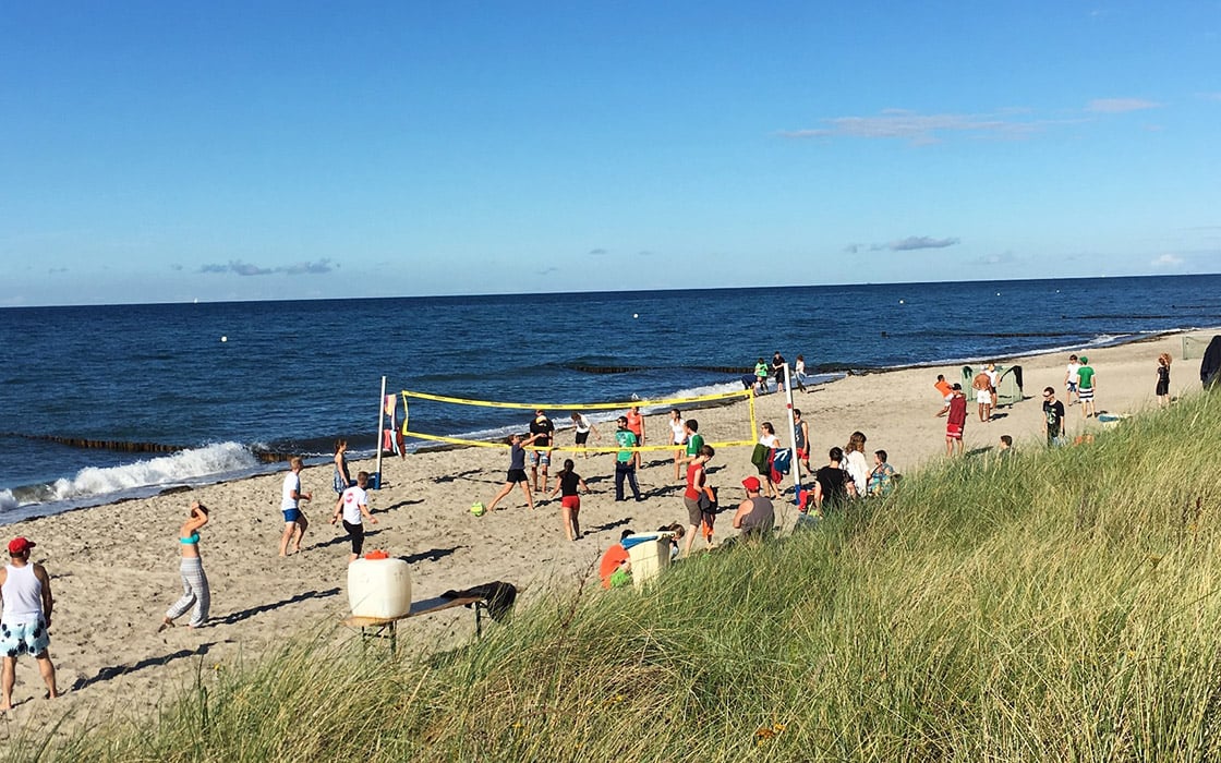 Menschen spielen Volleyball an einem Sandstrand in der Nähe des Ozeans, mit grasbewachsenen Dünen im Vordergrund unter einem klaren blauen Himmel.