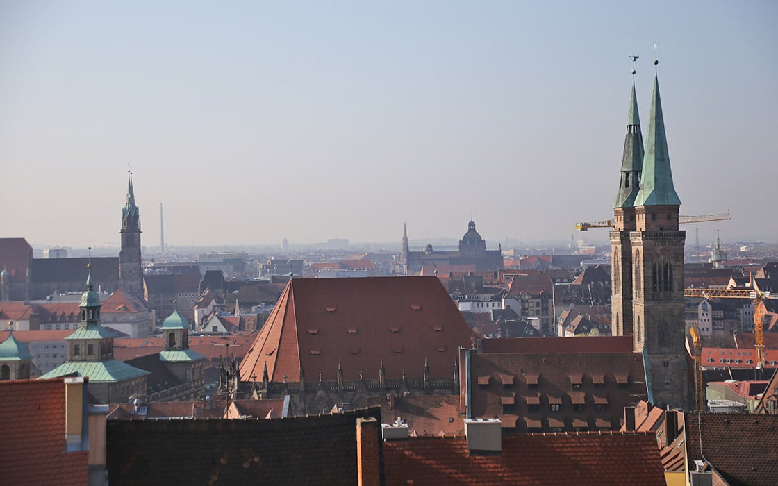 Stadtbild mit roten Dächern und Kirchtürmen unter einem dunstigen Himmel, wahrscheinlich in einer historischen europäischen Stadt.