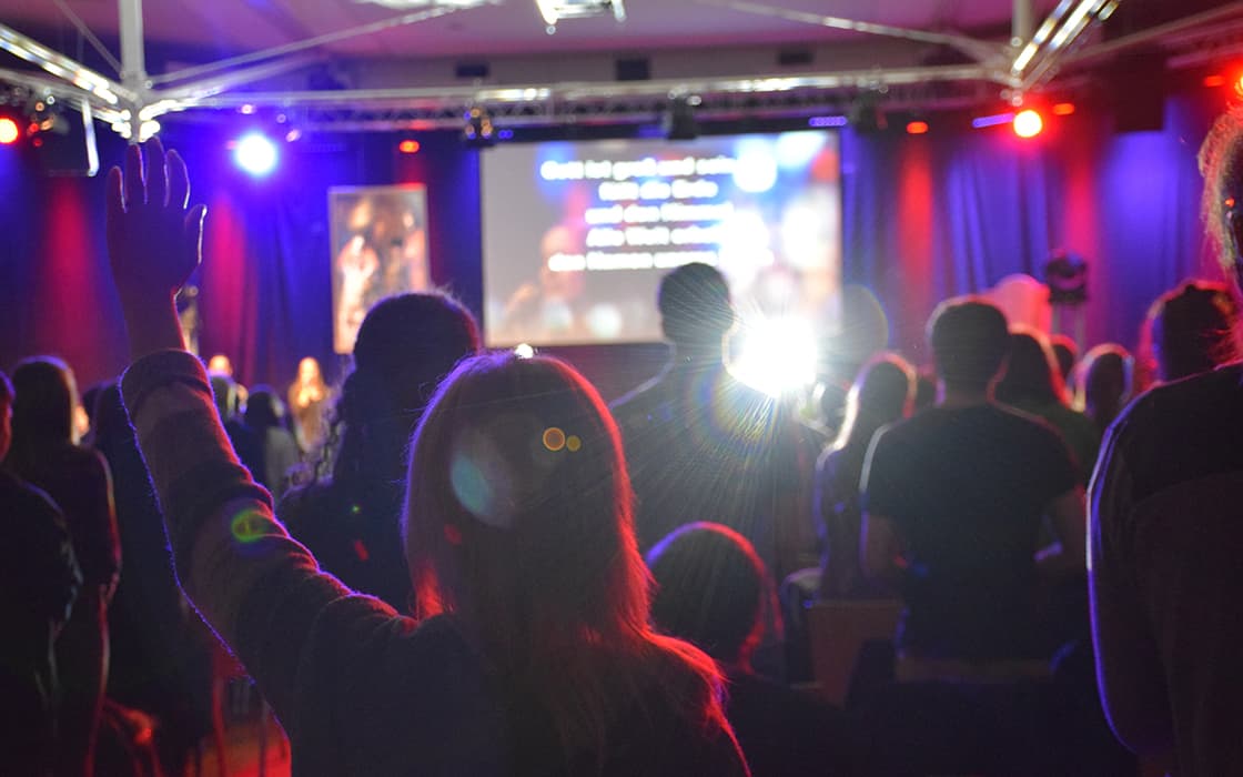 Publikum bei einem Konzert oder einer Veranstaltung mit bunten Bühnenlichtern, eine Person in der ersten Reihe hebt eine Hand und blickt zur Bühne.