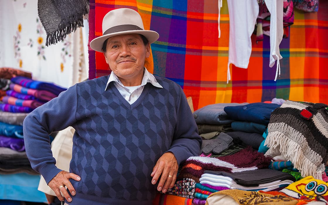 Ein lächelnder Mann mit Hut steht neben bunten, gewebten Textilien an einem Marktstand mit hellem Stoff im Hintergrund.