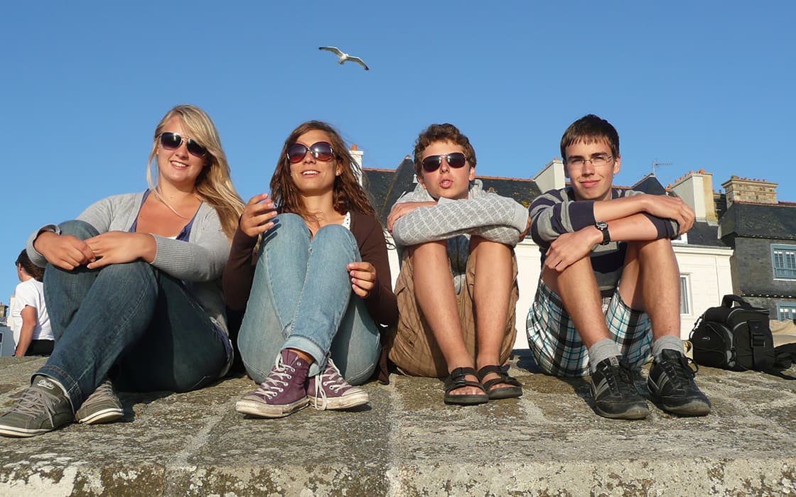 Vier junge Leute sitzen lächelnd auf einem Steinsims im Sonnenlicht, mit Häusern und einer fliegenden Möwe im Hintergrund.