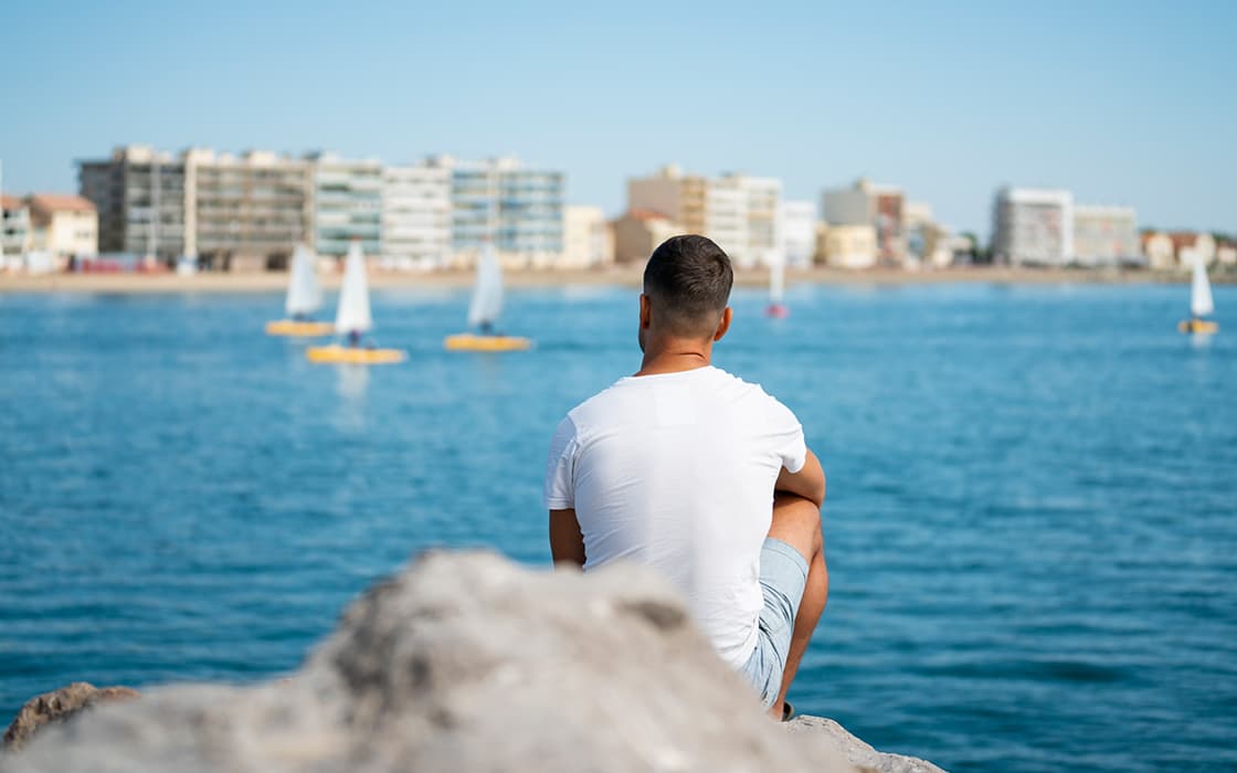Ein Mann in einem weißen Hemd sitzt auf einem Felsen und blickt auf das Meer hinaus, mit Segelbooten und Gebäuden im Hintergrund.