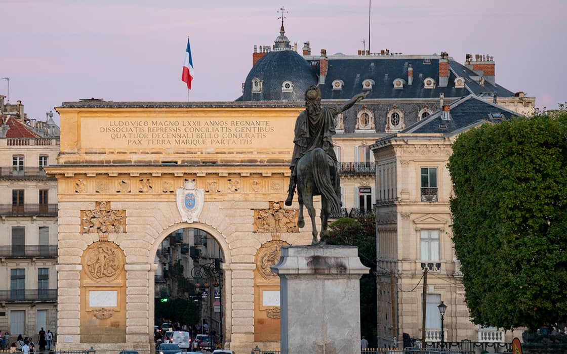 Eine Reiterstatue steht vor dem Arc de Triomphe in Montpellier, mit einer französischen Flagge auf dem Bogen bei Sonnenuntergang.