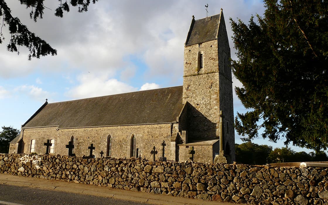 Alte Steinkirche mit hohem Turm und umliegendem Friedhof, begrenzt durch eine niedrige Steinmauer, unter einem bewölkten Himmel.