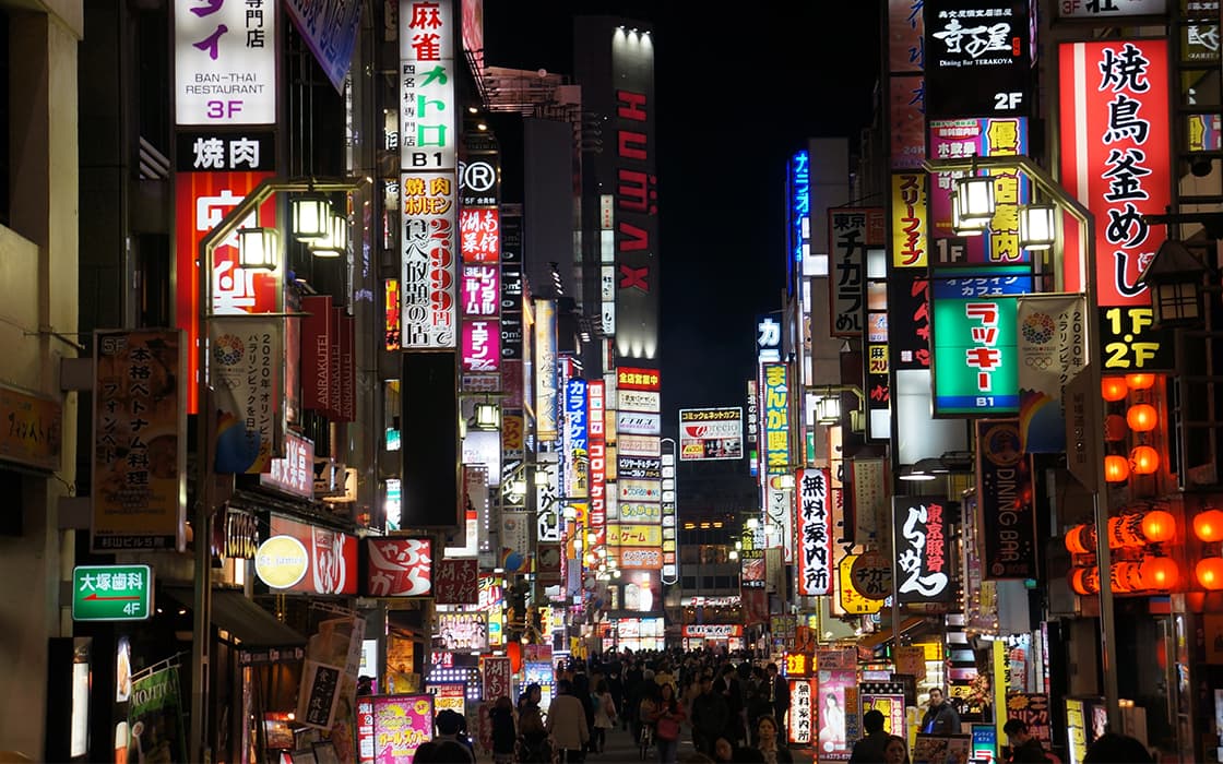Eine belebte nächtliche Straße in Japan, hell erleuchtet mit bunten Leuchtreklamen und Werbetafeln in japanischen Schriftzeichen.