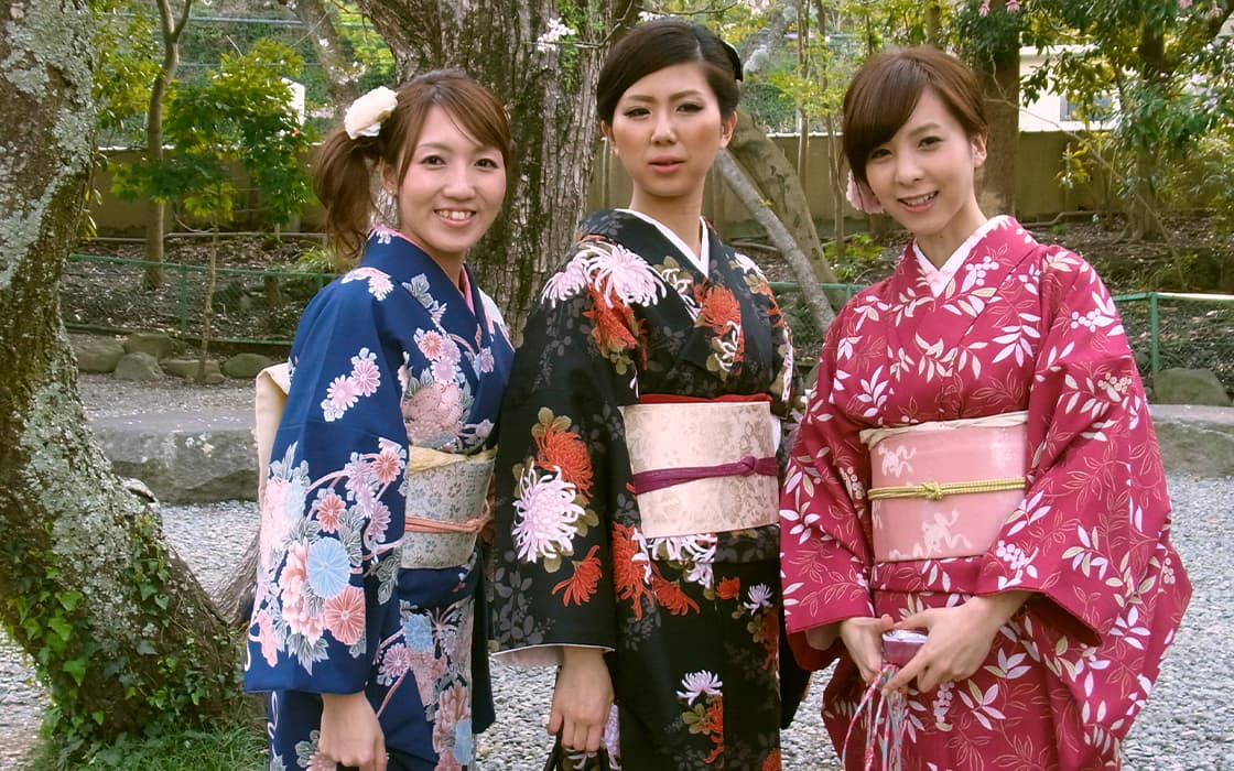 Drei Frauen in farbenfrohen, geblümten Kimonos stehen im Freien und lächeln zwischen Bäumen und Grünzeug.