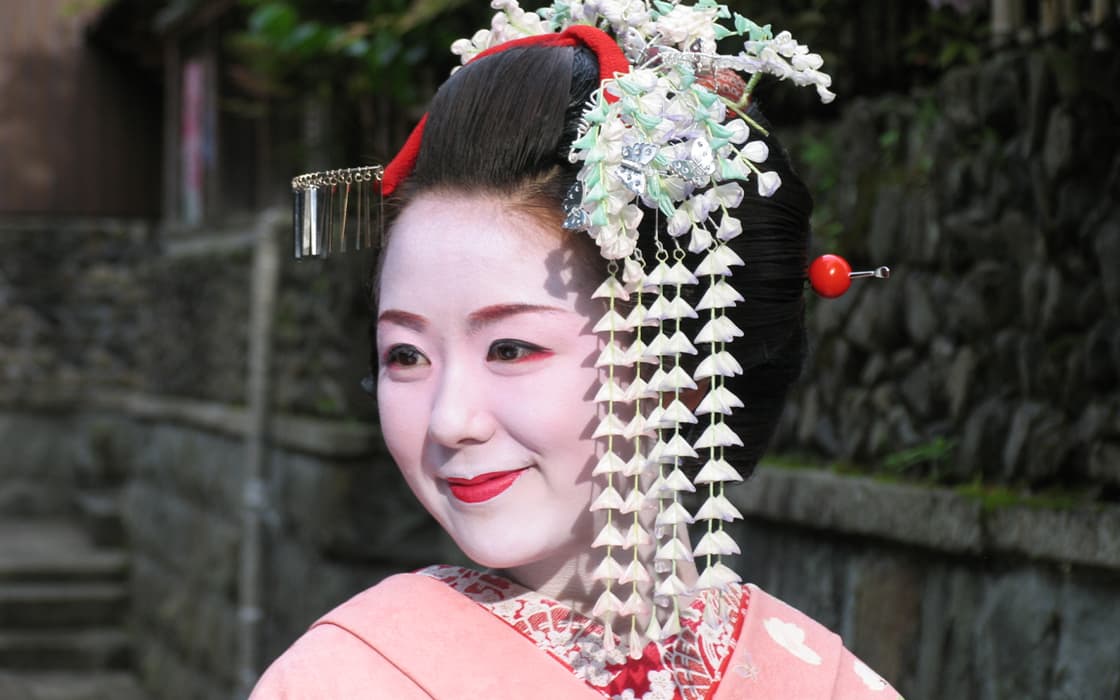 Eine Frau in traditioneller japanischer Geisha-Kleidung mit weißem Make-up und kunstvollem Haarschmuck, die im Freien lächelt.