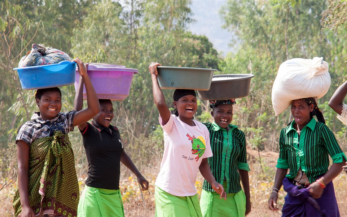 Fünf lächelnde Frauen gehen im Freien spazieren, jede trägt große Körbe oder Taschen auf dem Kopf, mit Bäumen im Hintergrund.