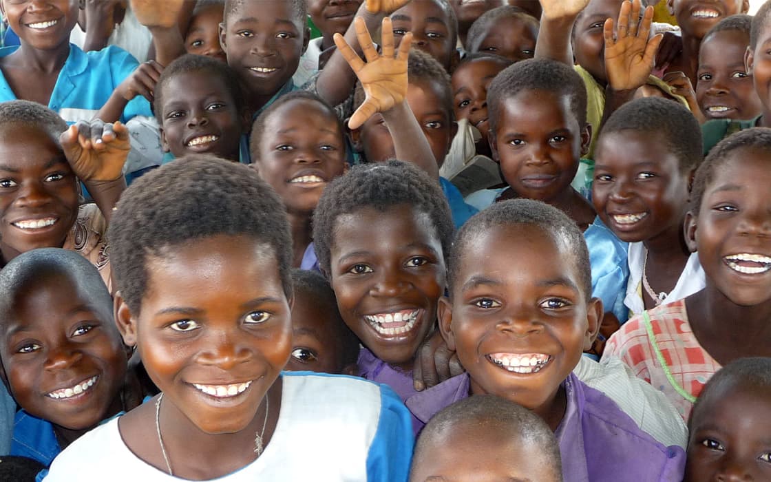 Eine Gruppe lächelnder Kinder, die dicht beieinander stehen, in die Kamera schauen und winken.