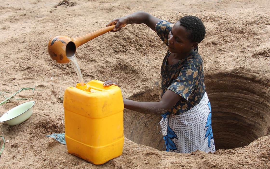 Frau holt Wasser aus einem flachen Brunnen und gießt es mit einer Schöpfkelle in einen gelben Behälter auf sandigem Boden.
