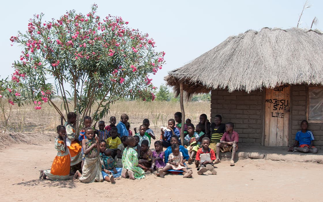 Kinder sitzen in Gruppen vor einer strohgedeckten Hütte und einem blühenden Baum in einer ländlichen Umgebung unter einem klaren Himmel.