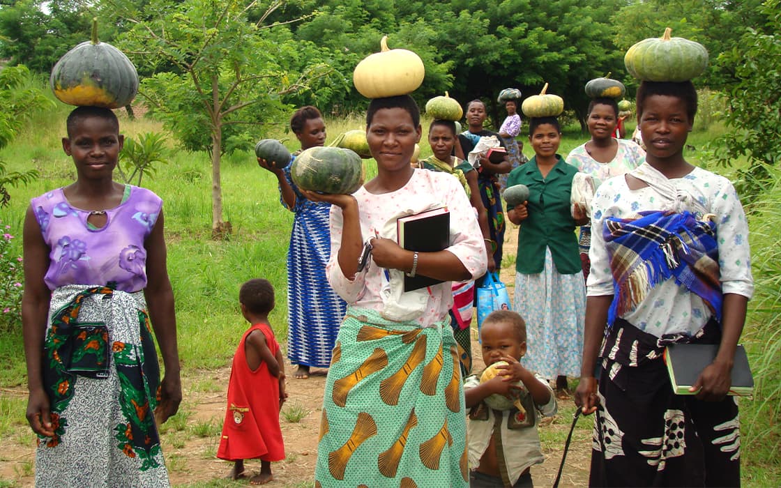 Eine Gruppe von Frauen und Kindern geht im Freien spazieren, einige Frauen balancieren einen Kürbis auf dem Kopf und halten Notizbücher in der Hand.