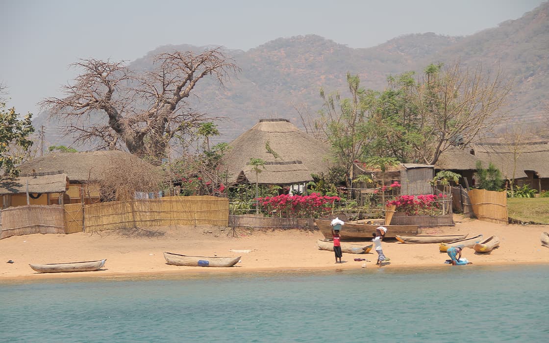 Strohgedeckte Hütten und Holzboote an einem sandigen Seeufer, mit Menschen und Bäumen, im Hintergrund die dunstigen Berge.