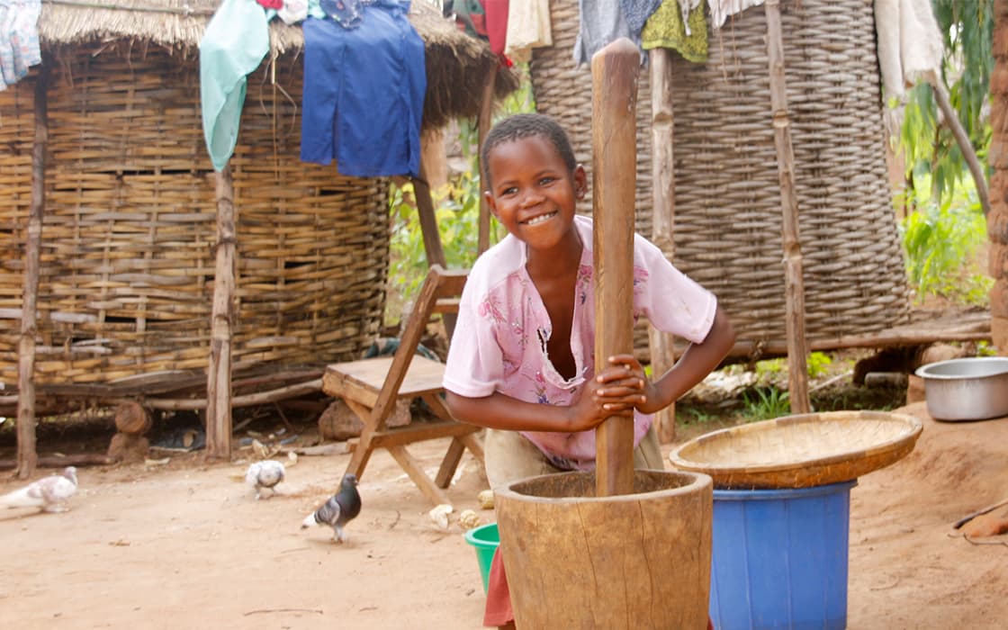 Lächelndes Kind in rosa Hemd benutzt einen großen hölzernen Stößel, um Lebensmittel im Freien zu zerstoßen, mit geflochtenen Hütten und Wäsche im Hintergrund.