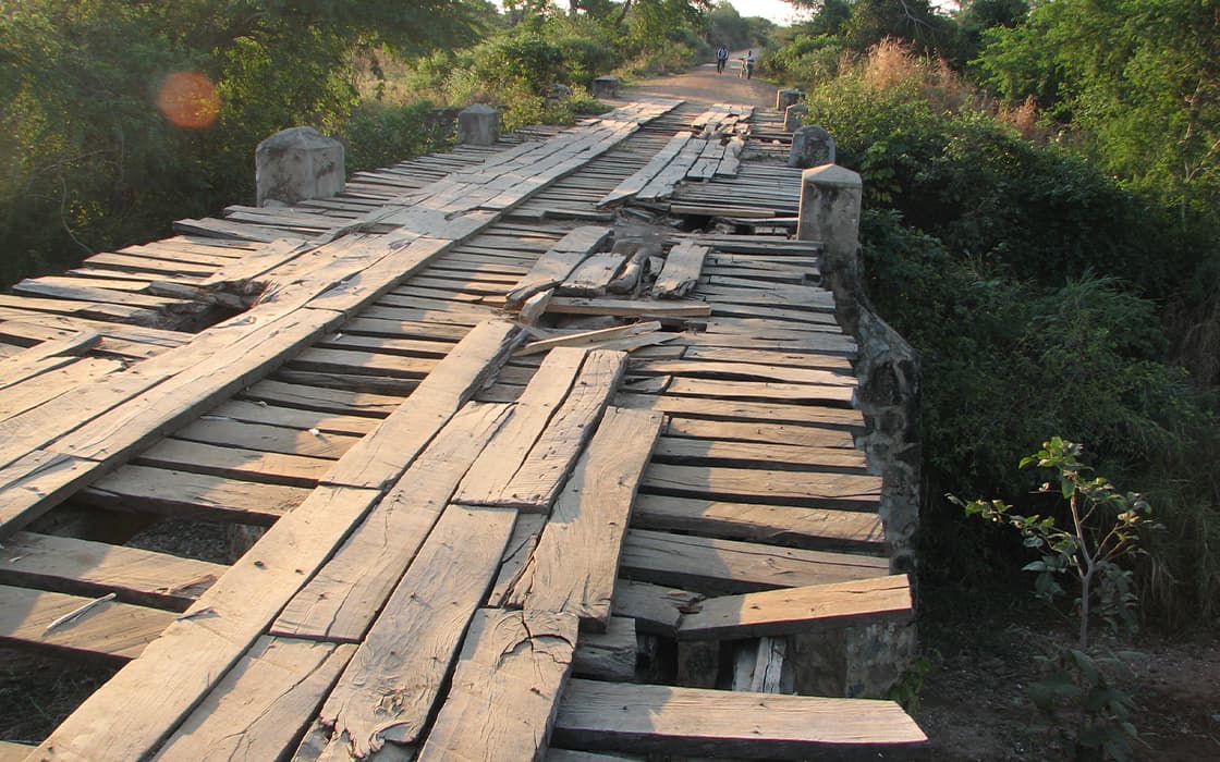 Eine beschädigte Holzbrücke mit fehlenden und unebenen Bohlen überquert überwuchertes Land im Sonnenlicht.