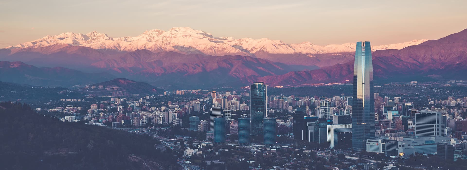 Skyline der Stadt mit hohen modernen Gebäuden, Bergen im Hintergrund und schneebedeckten Gipfeln bei Sonnenuntergang.