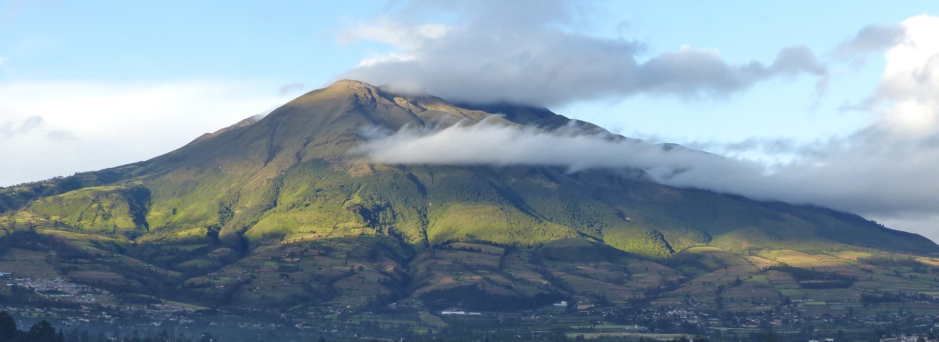 Ein grüner Berg mit Wolkenflecken auf dem Gipfel und in der Mitte unter einem teilweise bewölkten Himmel.
