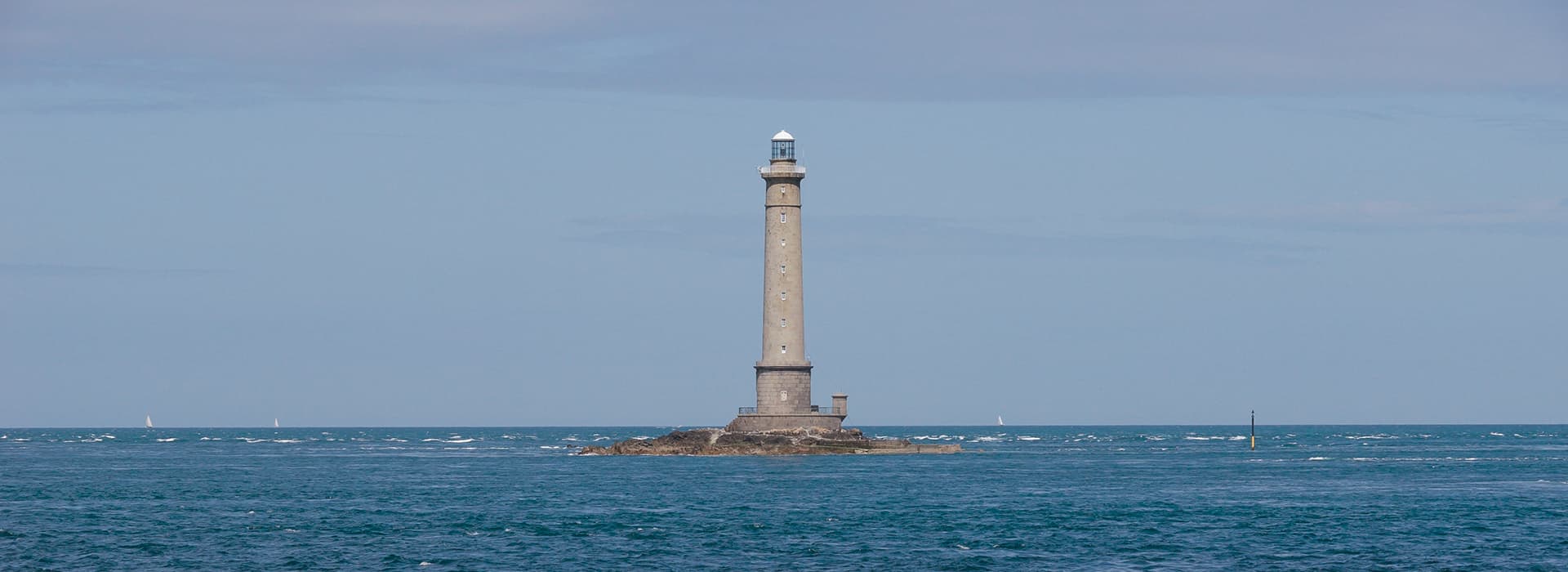 Ein hoher Leuchtturm steht auf einer kleinen felsigen Insel, umgeben von blauem Meer und einem teilweise bewölkten Himmel.
