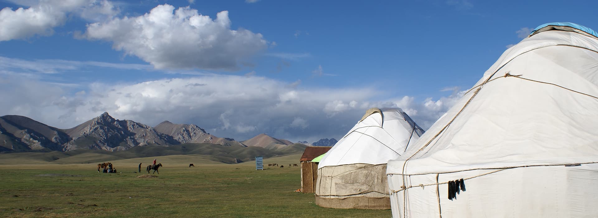 Weiße Jurten auf grasbewachsenen Ebenen mit fernen Bergen, unter einem blauen Himmel mit vereinzelten Wolken. Menschen und Pferde im Hintergrund.