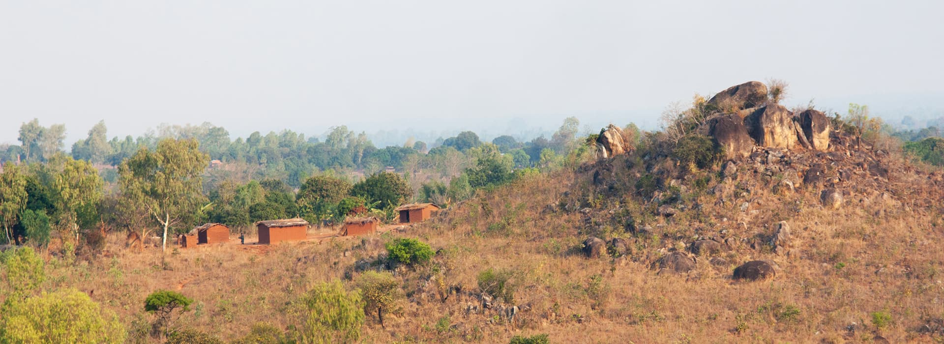 Kleine Backsteinhäuser und vereinzelte Bäume stehen in der Nähe eines felsigen Hügels in einer trockenen, ländlichen Landschaft unter einem dunstigen Himmel.