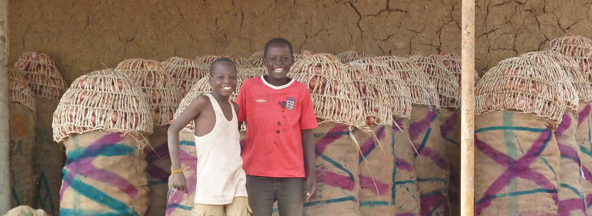 Zwei lächelnde Jungen stehen vor Säcken mit Waren, die mit geflochtenen Körben bedeckt sind, vor dem Hintergrund einer Lehmwand.