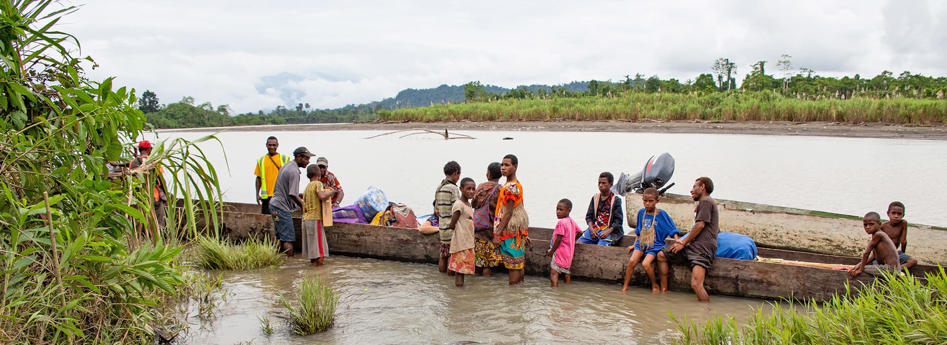 Eine Gruppe von Menschen in Papua-Neuguinea steht an einem Flussufer mit Holzkanus, umgeben von üppigem Grün.