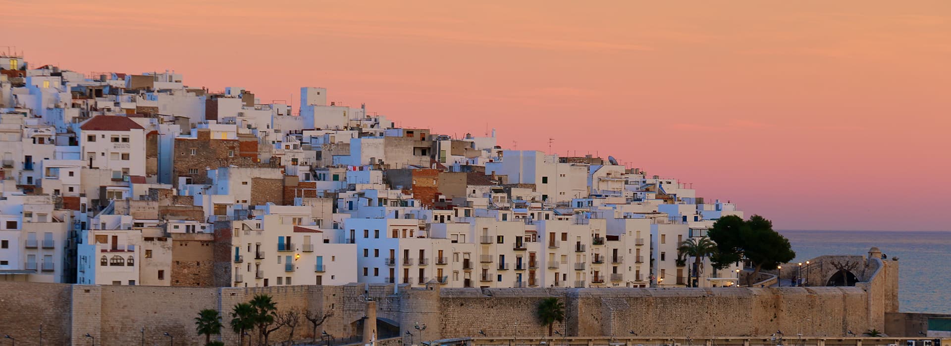 Eine Ansammlung weißer Gebäude auf einem Hügel am Meer bei Sonnenuntergang, mit einem rosa und orangefarbenen Himmel im Hintergrund.