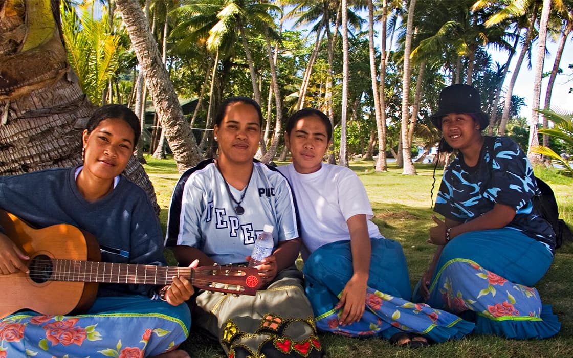 Vier Frauen sitzen im Gras unter Palmen; eine von ihnen spielt auf einer Gitarre. Sie tragen alle geblümte Röcke und posieren für das Foto.