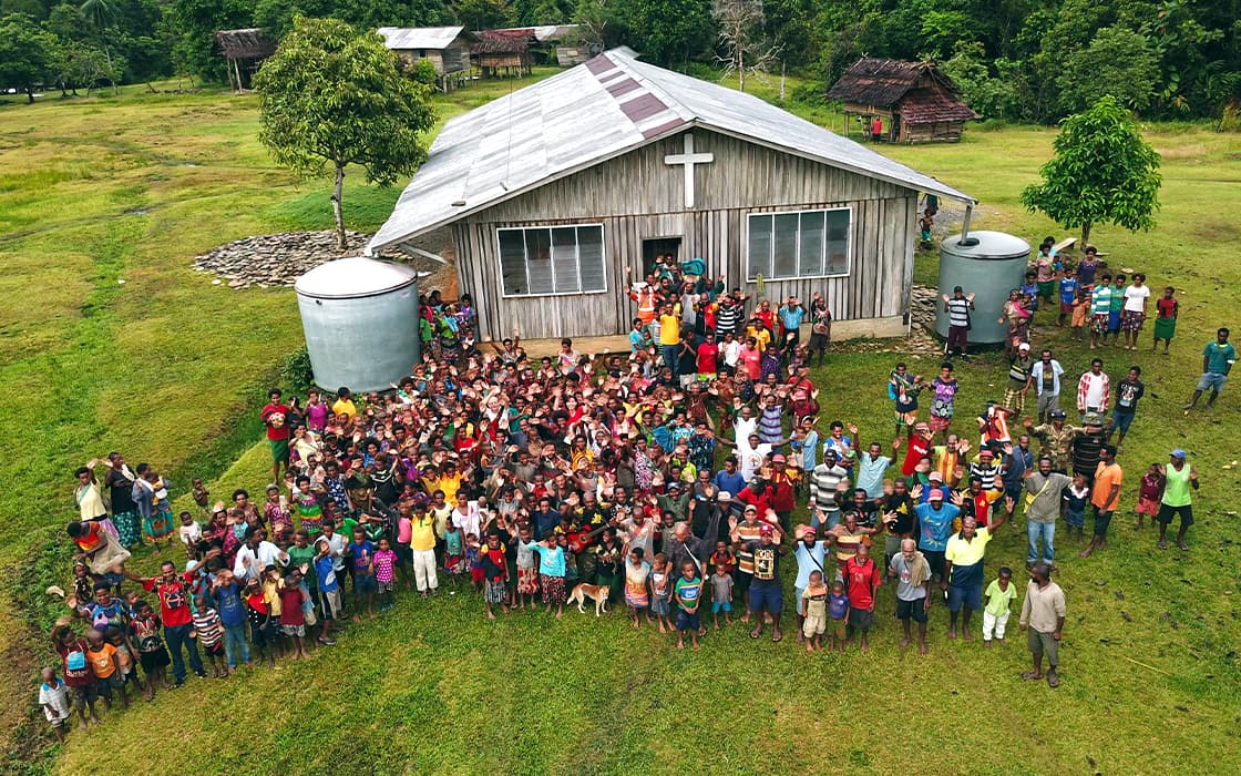 Eine große Gruppe versammelt sich vor einer Holzkirche mit einem Kreuz im ländlichen, grünen Papua-Neuguinea.