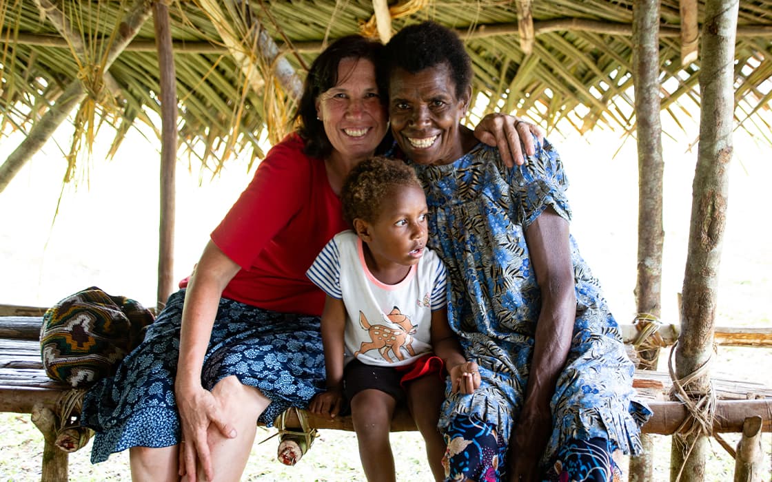 Drei Personen sitzen auf einer Holzbank unter einem Strohdach in Papua-Neuguinea, zwei Frauen und ein lächelndes Kind.
