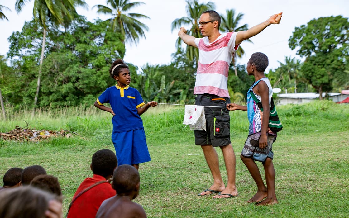 In Papua-Neuguinea steht ein Mann mit weit ausgebreiteten Armen, während zwei Kinder auf einer Wiese eine Schnur halten.