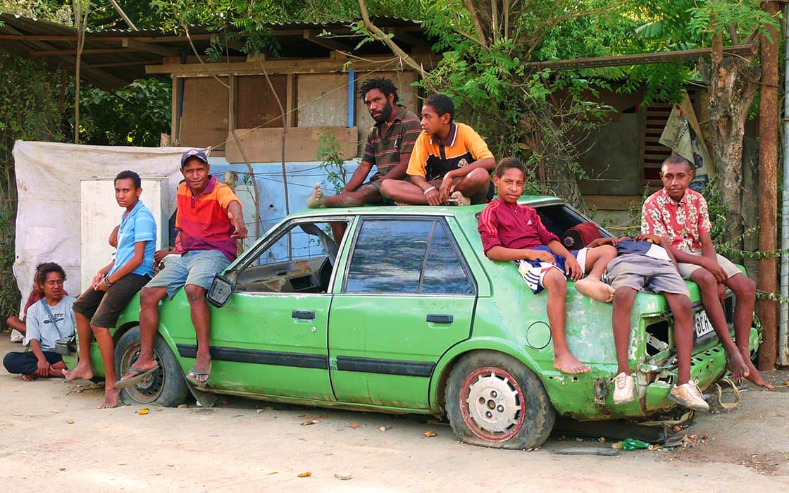Eine Gruppe faulenzt auf einem abgenutzten, grünen Auto in Papua-Neuguinea, das in der Nähe von Bäumen und einem rustikalen Gebäude geparkt ist.