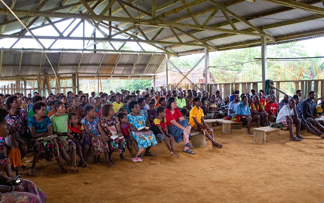 Eine große Gruppe von Menschen sitzt auf Bänken in einem offenen Gebäude in Papua-Neuguinea mit einem Wellblechdach.