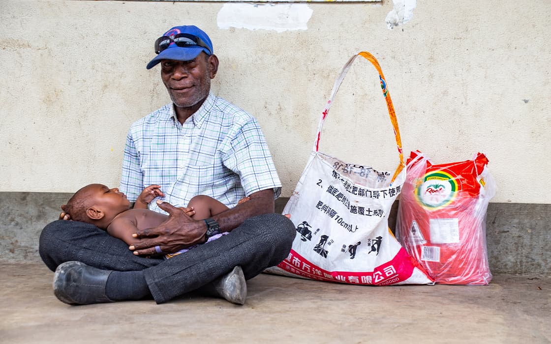 Ein älterer Mann in Papua-Neuguinea sitzt im Schneidersitz und füttert ein Baby, während er Säcke mit Lebensmitteln an einer Wand lehnt.