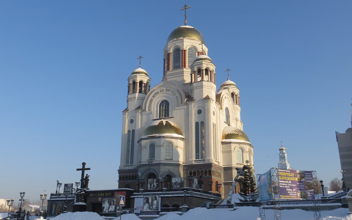 Eine große weiße Kirche mit goldenen Kuppeln steht unter einem klaren blauen Himmel, umgeben von Schnee und mehreren Denkmälern.